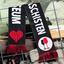 Lade das Bild in den Galerie-Viewer, Das Bild zeigt einen Schal der über einem Zaun hängt. Er ist schwarz und hat schwarze und rote Fransen. Es ist ein Kreislogo darauf mit weißem Hintergrund und einem schwarzen und einem roten Weinglas. In Anlehnung an das klassische Antifa Logo. Ebenso ist ein rotes Herz darauf, welches aus den Zahlen 1910 das Herz bildet. Man erkennt auch große weiße Buchstaben. Diese ergeben auf der einen Seite den Satz Kein Wein den Faschisten und auf der anderen Seite FC St. Pauli Museum.