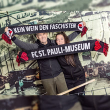Lade das Bild in den Galerie-Viewer, Auf dem Bild sieht man 2 Models vor einer Wand. Auf der ist eine Szene der alten Hafenstraße abgebildet. Die 2 Models halten jeweils einen Schal hoch. Er ist schwarz und es steht in großen Buchstaben der Satz Kein Wein den Faschisten darauf. Auf der anderen Rückseite des Schals steht in großen weißen Buchstaben FC St. Pauli Museum.