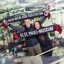 Lade das Bild in den Galerie-Viewer, Auf dem Bild sieht man 2 Models vor einer Wand. Auf der ist eine Szene der alten Hafenstraße abgebildet. Die 2 Models halten jeweils einen Schal hoch. Er ist schwarz und es steht in großen Buchstaben der Satz Kein Wein den Faschisten darauf. Auf der anderen Rückseite des Schals steht in großen weißen Buchstaben FC St. Pauli Museum.