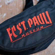 Lade das Bild in den Galerie-Viewer, Auf dem Bild sieht man eine Nahaufnahme einer schwarzen Gürteltasche. FC St. Pauli Museum ist in rot darauf eingestickt.
