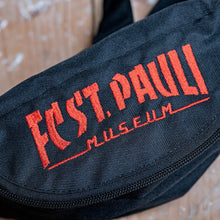 Lade das Bild in den Galerie-Viewer, Auf dem Bild sieht man eine Nahaufnahme einer schwarzen Gürteltasche. FC St. Pauli Museum ist in rot darauf eingestickt.