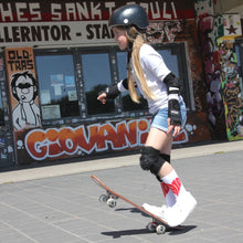 Lade das Bild in den Galerie-Viewer, Auf dem Foto sieht man ein Mädchen mit Skateboard. Sie trägt einen schwarzen Helm schwarze Knie Handgelenk und Ellbogen Schützer. Außerdem ein weißes T Shirt eine blaue kurze Jeans sowie weiße Socken mit einem großen roten 1910 Herz darauf. Im Hintergrund erkennt man die Fanräume in der Gegengerade des Millerntor Stadions. Heimat des FC St.Pauli.