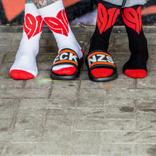 Lade das Bild in den Galerie-Viewer, Auf dem Foto sieht man die Füße zweier Models. Das linke Model trägt weiße Socken bei denen Fersen und Zehenbereich rot sind sowie jeweils mittig groß ein rotes 1910 Herz eingestickt ist. Das rechte Model trägt schwarze Socken bei denen Fersen und Zehenbereich rot sind und jeweils mittig groß ein rotes 1910 Herz eingestickt ist. Die Models stehen überkreuzt da und tragen jeweils einen Badelatschen. Zusammen nebeneinander ergeben die schwarz weiß roten Badelatschen die Wörter FCK NZS.
