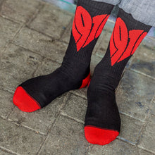 Lade das Bild in den Galerie-Viewer, Auf dem Foto sieht man 2 Füße eines Models. Das Model steht auf einem Steinboden und trägt schwarze Socken mit einem großen, roten 1910 Herz eingestickt. Zehenbereich und Ferse sind ebenfalls rot.