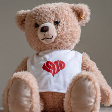Lade das Bild in den Galerie-Viewer, Auf dem Bild sieht man einen hellbraunen Teddybären. Dieser trägt ein weißes Lätzchen mit dem roten Aufdruck 1910. Die Zahlen sind herzförmig angeordnet.