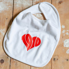 Lade das Bild in den Galerie-Viewer, Auf dem Bild sieht man einen Holzfußboden. Auf diesem liegt ein weißer Babylatz mit rotem Aufdruck 1910. Die Zahlen sind herzförmig angeordnet.