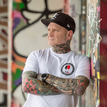 Lade das Bild in den Galerie-Viewer, Ihr seht auf dem Foto ein Model angelehnt an eine bunte Wand. Er trägt ein weißes T Shirt mit dem Pocketprint Kein Wein den Faschisten sowie 1910 Weinbar St.Pauli und einem schwarzen und einem roten Weinglas. In Anlehnung an das klassische Antifa Logo. Das Bild entstand vor der Gegengerade des Millerntor Stadions. Heimat des FC St.Pauli.