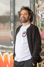 Lade das Bild in den Galerie-Viewer, Ihr seht auf dem Foto ein Model vor einer bunten Wand. Er trägt ein weißes T Shirt mit dem Pocketprint Kein Wein den Faschisten sowie 1910 Weinbar St.Pauli und einem schwarzen und einem roten Weinglas. In Anlehnung an das klassische Antifa Logo. Das Bild entstand vor der Gegengerade des Millerntor Stadions. Heimat des FC St.Pauli.