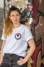 Lade das Bild in den Galerie-Viewer, Ihr seht auf dem Foto ein Model angelehnt an eine bunte Wand. Sie trägt ein weißes T Shirt mit dem Pocketprint Kein Wein den Faschisten sowie 1910 Weinbar St.Pauli und einem schwarzen und einem roten Weinglas. In Anlehnung an das klassische Antifa Logo. Das Bild entstand vor der Gegengerade des Millerntor Stadions. Heimat des FC St.Pauli.