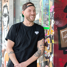 Lade das Bild in den Galerie-Viewer, Auf dem Bild sieht man ein Model an eine bunte Wand gelehnt. Er trägt ein schwarzes T Shirt mit kleinem weißen 1910 Herz auf der linken Seite aufgestickt. Das Foto entstand vor der Gegengerade des Millerntor Stadions. Heimat des FC St.Pauli.