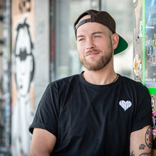 Lade das Bild in den Galerie-Viewer, Auf dem Bild sieht man ein Model an eine bunte Wand gelehnt. Er trägt ein schwarzes T Shirt mit kleinem weißen 1910 Herz auf der linken Seite aufgestickt. Das Foto entstand vor der Gegengerade des Millerntor Stadions. Heimat des FC St.Pauli.