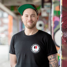 Lade das Bild in den Galerie-Viewer, Auf dem Bild steht 1 Model vor bunten Wänden. Er trägt ein schwarzes Shirt mit einem runden Pocketprint auf der linken Seite. Auf diesem steht Kein Wein den Faschisten 1910 Weinbar St.Pauli und in der Mitte sind 2 Weingläser. Ein rotes und ein schwarzes. In Anlehnung an das klassische Antifa Logo. Das Foto entstand vor der Gegengerade des Millerntor Stadions. Heimat des FC St. Pauli.