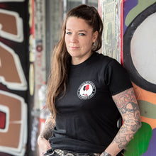 Lade das Bild in den Galerie-Viewer, Auf dem Bild steht 1 Model angelehnt an eine bunte Wand. Sie trägt ein schwarzes Shirt mit einem runden Pocketprint auf der linken Seite. Auf diesem steht Kein Wein den Faschisten 1910 Weinbar St.Pauli und in der Mitte sind 2 Weingläser. Ein rotes und ein schwarzes. In Anlehnung an das klassische Antifa Logo. Das Foto entstand vor der Gegengerade des Millerntor Stadions. Heimat des FC St. Pauli.