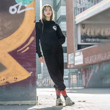 Lade das Bild in den Galerie-Viewer, Ihr seht auf dem Bild 1 Model angelehnt an eine bunte Wand. Sie trägt einen schwarzen Kapuzenpulli mit dem Pocketprint Kein Wein den Faschisten sowie 1910 Weinbar St.Pauli und einem roten und einem schwarzen Weinglas. In Anlehnung an das klassische Antifa Logo. Das Foto entstand vor der Gegengerade des Millerntor Stadions. Heimat des FC St.Pauli.