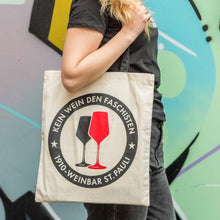 Lade das Bild in den Galerie-Viewer, Auf dem Bild sieht man einen naturfarbenen Jutebeutel mit schwarzen Henkeln. Auf der Tasche befindet sich der Aufdruck Kein Wein den Faschisten sowie 1910 Weinbar St.Pauli und einem schwarzen und einem roten Weinglas. In Anlehnung an das klassische Antifa Logo.