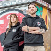 Lade das Bild in den Galerie-Viewer, Ihr seht auf dem Bild 2 Models vor einer bunten Wand. Sie tragen schwarze Kapuzenpullis mit dem Pocketprint Kein Wein den Faschisten sowie 1910 Weinbar St.Pauli und einem roten und einem schwarzen Weinglas. In Anlehnung an das klassische Antifa Logo. Das Foto entstand vor dem Millerntor Stadion. Heimat des FC St.Pauli.