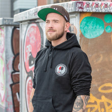 Lade das Bild in den Galerie-Viewer, Ihr seht auf dem Bild 1 Model vor einer bunten Wand. Er trägt einen schwarzen Kapuzenpulli mit dem Pocketprint Kein Wein den Faschisten sowie 1910 Weinbar St.Pauli und einem roten und einem schwarzen Weinglas. In Anlehnung an das klassische Antifa Logo. Das Foto entstand vor dem Millerntor Stadion. Heimat des FC St.Pauli.