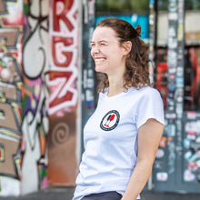 Lade das Bild in den Galerie-Viewer, Auf dem Foto sieht man ein Model vor einer bunten Wand. Sie trägt ein tailliertes, weißes Shirt mit dem Aufdruck Kein Wein den Faschisten sowie 1910 Weinbar St.Pauli und einem schwarzen und einem roten Weinglas. In Anlehnung an das klassische Antifa Logo. Das Bild entstand vor der Gegengerade des Millerntor Stadions. Heimat des FC St.Pauli.