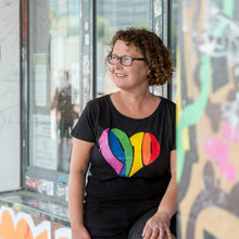 Lade das Bild in den Galerie-Viewer, Auf dem Foto sieht man ein Model an eine bunte Wand gelehnt. Sie trägt ein schwarzes tailliertes Shirt. Auf dem Shirt ist ein großes 1910 Herz in regenbogenfarben gedruckt. Das Foto entstand vor dem Millerntor Stadion. Heimat des FC St.Pauli.