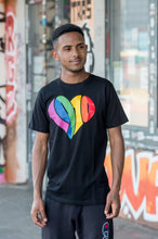 Lade das Bild in den Galerie-Viewer, Auf dem Bild sieht man ein Model in schwarzem T Shirt mit einem großen 1910 Herz Druck in regenbogenfarben. Das Model steht vor einer bunten Wand. Das Foto entstand vor dem Millerntor Stadion. Heimat des FC St.Pauli.
