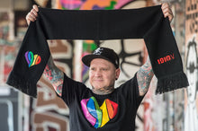 Lade das Bild in den Galerie-Viewer, Auf dem Foto sieht man ein Model der mit beiden Armen einen Wollschal nach oben hält. Auf dem Schal ist an einem Ende ein regenbogenfarbenes 1910 Herz gestickt. Am anderen Ende ist in rot 1910 eV gestickt. Das Model trägt außerdem ein schwarzes Shirt auf dem mittig ein großes 1910 Herz in regenbogenfarben gedruckt ist. Das Foto entstand vor dem Millerntorstadion. Heimat des FC St.Pauli.