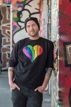 Lade das Bild in den Galerie-Viewer, Auf dem Foto sieht man ein Model an eine bunte Wand gelehnt. Er trägt eine schwarze Wollmütze die unten umgeschlagen ist und darauf ist ein regenbogenfarbenes 1910 Herz gestickt. Außerdem trägt er ein schwarzes Sweatshirt mit einem großen 1910 Herz in regenbogenfarben gedruckt. Das Foto wurde vor dem Millerntor Stadion aufgenommen. Heimat des FC St.Pauli.