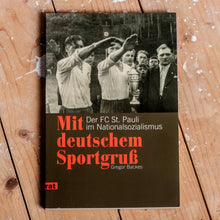 Lade das Bild in den Galerie-Viewer, Auf dem Bild sieht man einen Holzfußboden auf dem ein Buch liegt. In der oberen Hälfte des Buches sieht man ein altes Foto mit Sportlern aus der NS Zeit die den rechten Arm heben. Darunter befindet sich der Schriftzug Mit deutschem Sportgruß Der FC St.Pauli im Nationalsozialismus sowie der Name des Autors Gregor Backes