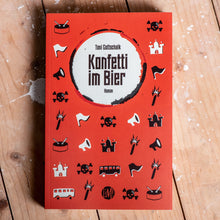Lade das Bild in den Galerie-Viewer, Auf dem Bild sieht man einen Holzfußboden. Auf diesem liegt ein rotes Buch auf dem steht Toni Gottschalk Konfetti im Bier Roman. Es befinden sich außerdem viele, kleine, verschiedene Grafiken auf dem Cover. Diese sind schwarz und weiß. Zum Beispiel Totenköpfe, Busse, Trommeln, Fahnen und mehr.