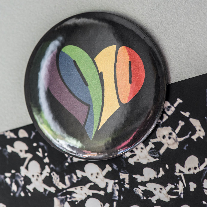 Auf dem Bild sieht man einen runden Magnet. Die Grundfarbe ist schwarz und es ist ein 1910 Herz in regenbogenfarben aufgedruckt.