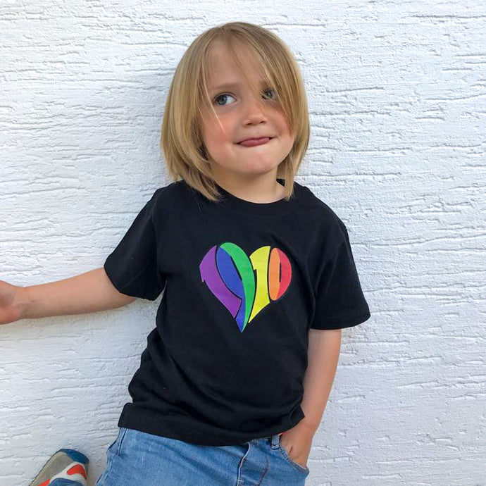 Auf dem Foto sieht man ein Kind vor einer weißen Wand. Es trägt eine blaue Jeans und ein schwarzes T Shirt mit 1910 Herz in regenbogenfarben aufgedruckt.