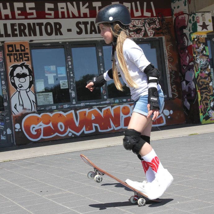 Auf dem Foto sieht man ein Mädchen mit Skateboard. Sie trägt einen schwarzen Helm schwarze Knie Handgelenk und Ellbogen Schützer. Außerdem ein weißes T Shirt eine blaue kurze Jeans sowie weiße Socken mit einem großen roten 1910 Herz darauf. Im Hintergrund erkennt man die Fanräume in der Gegengerade des Millerntor Stadions. Heimat des FC St.Pauli.