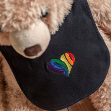 Lade das Bild in den Galerie-Viewer, Auf dem Bild sieht man einen hellbraunen Teddybären. Dieser trägt einen schwarzen Babylatz. Auf dem Lätzchen ist mittig ein 1910 Herz in regenbogenfarben eingestickt.

