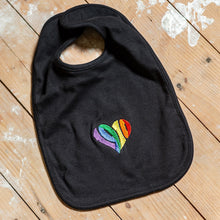 Lade das Bild in den Galerie-Viewer, Auf dem Bild sieht man einen Holzfußboden. Auf dem Boden liegt ein schwarzer Babylatz auf dem mittig ein regenbogenfarbenes 1910 Herz eingestickt ist.
