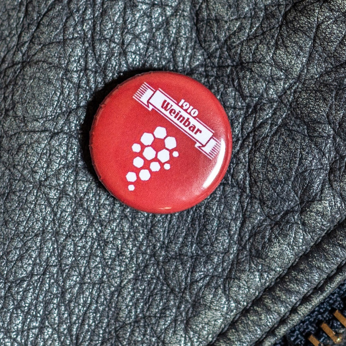 Auf dem Foto sieht man einen roten Button mit dem weißen Aufdruck 1910 Weinbar und einer Traube in Fußball Optik. Der Button befindet sich auf einer Lederjacke.