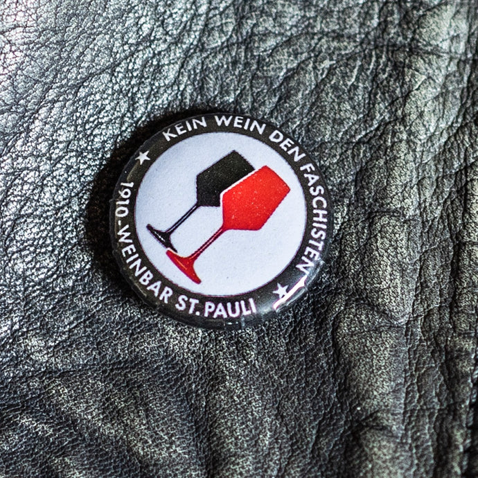 Auf dem Foto sieht man einen Button mit dem Aufdruck Kein Wein den Faschisten sowie 1910 Weinbar St.Pauli und einem schwarzen und einem roten Weinglas. In Anlehnung an das klassische Antifa Logo. Der Button befindet sich auf einer Lederjacke.