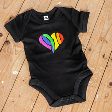 Lade das Bild in den Galerie-Viewer, Auf dem Bild sieht man einen Holzfußboden. Auf diesem liegt ein schwarzer Babybody mit einem regenbogenfarbenen 1910 Herz Aufdruck.
