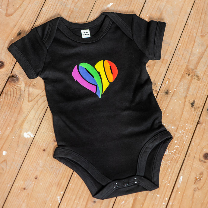 Auf dem Bild sieht man einen Holzfußboden. Auf diesem liegt ein schwarzer Babybody mit einem regenbogenfarbenen 1910 Herz Aufdruck.