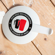 Auf dem Bild sieht man einen Holzfußboden. Auf diesem liegt ein weißer runder Becherdeckel. Auf diesem ist der Aufdruck Kein Bier den Faschisten zu sehen. Sowie 1910 Weinbar St.Pauli und 2 Stadionbecher. Ein schwarzer und ein roter. Angelehnt an das klassische Antifa Logo.