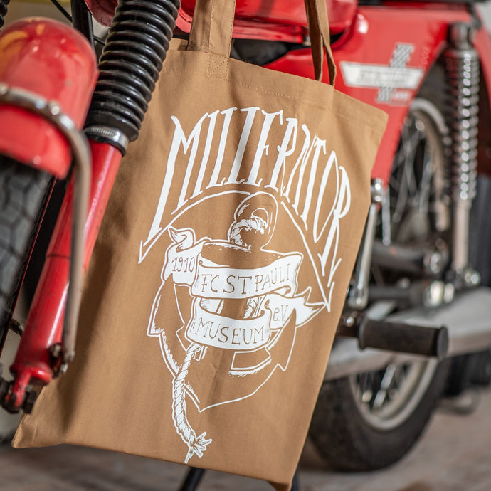 Eine braune Stofftasche hängt an einem roten Motorrad. Auf dem Stoffbeutel ist ein Ankermotiv in weiß. Darüber steht groß der Schriftzug Millermtor. Auf einem Spruchband, das um den Anker gewickelt ist steht: 1910 Fc St. pauli Museum e.V.