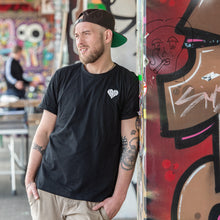 Lade das Bild in den Galerie-Viewer, Auf dem Bild sieht man ein Model an eine bunte Wand gelehnt. Er trägt ein schwarzes T Shirt mit kleinem weißen 1910 Herz auf der linken Seite aufgestickt. Das Foto entstand vor der Gegengerade des Millerntor Stadions. Heimat des FC St.Pauli.
