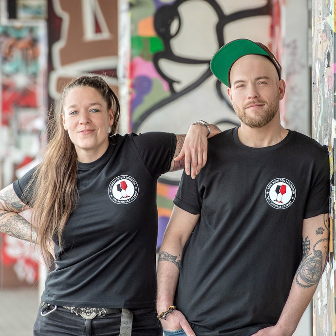 Auf dem Bild sind 2 Models vor einer bunten Wand. Sie tragen schwarze Shirts mit einem runden Pocketprint auf der linken Seite. Auf diesem steht Kein Wein den Faschisten 1910 Weinbar St.Pauli und in der Mitte sind 2 Weingläser. Ein rotes und ein schwarzes. In Anlehnung an das klassische Antifa Logo. Das Foto entstand vor der Gegengerade des Millerntor Stadions. Heimat des FC St. Pauli.