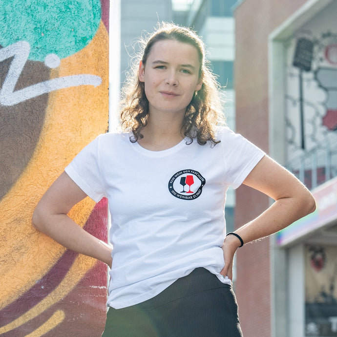 Auf dem Foto sieht man ein Model vor einer bunten Wand. Sie trägt ein tailliertes, weißes Shirt mit dem Aufdruck Kein Wein den Faschisten sowie 1910 Weinbar St.Pauli und einem schwarzen und einem roten Weinglas. In Anlehnung an das klassische Antifa Logo. Das Bild entstand vor der Gegengerade des Millerntor Stadions. Heimat des FC St.Pauli. 