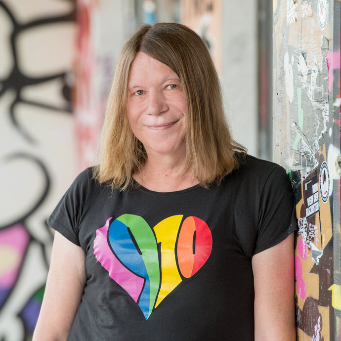 Auf dem Foto sieht man ein Model an eine bunte Wand gelehnt. Sie trägt ein schwarzes tailliertes Shirt. Auf dem Shirt ist ein großes 1910 Herz in regenbogenfarben gedruckt. Das Foto entstand vor dem Millerntor Stadion. Heimat des FC St.Pauli.