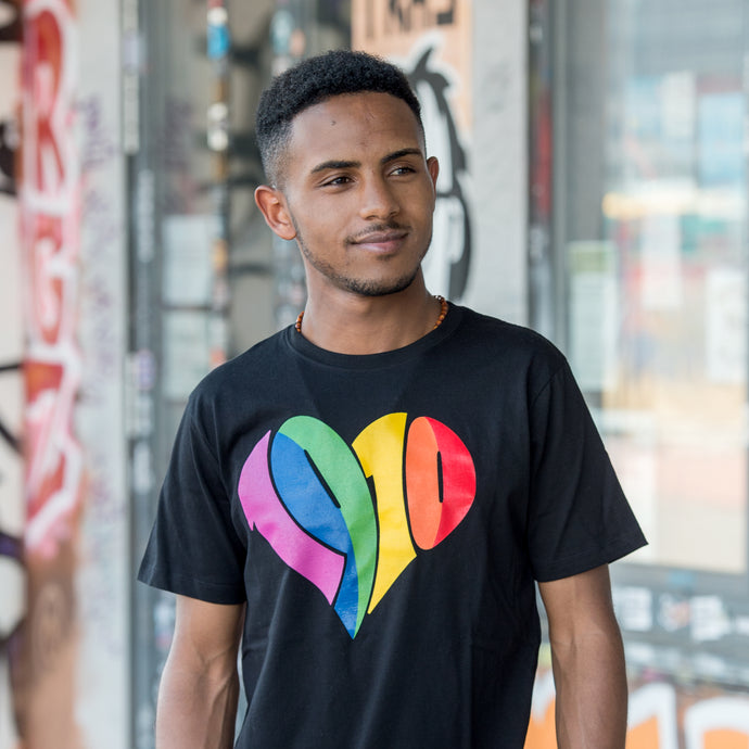 Auf dem Bild sieht man ein Model in schwarzem T Shirt mit einem großen 1910 Herz Druck in regenbogenfarben. Das Model steht vor einer bunten Wand. Das Foto entstand vor dem Millerntor Stadion. Heimat des FC St.Pauli.