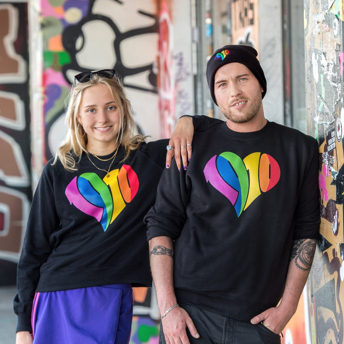 Auf dem Foto sieht man zwei Models an eine bunte Wand gelehnt. Sie tragen jeweils ein schwarzes Sweatshirt mit großem regenbogenfarbenen 1910 Herz Druck. Das Foto wurde vor dem Millerntor Stadion aufgenommen. Heimat des FC St.Pauli.