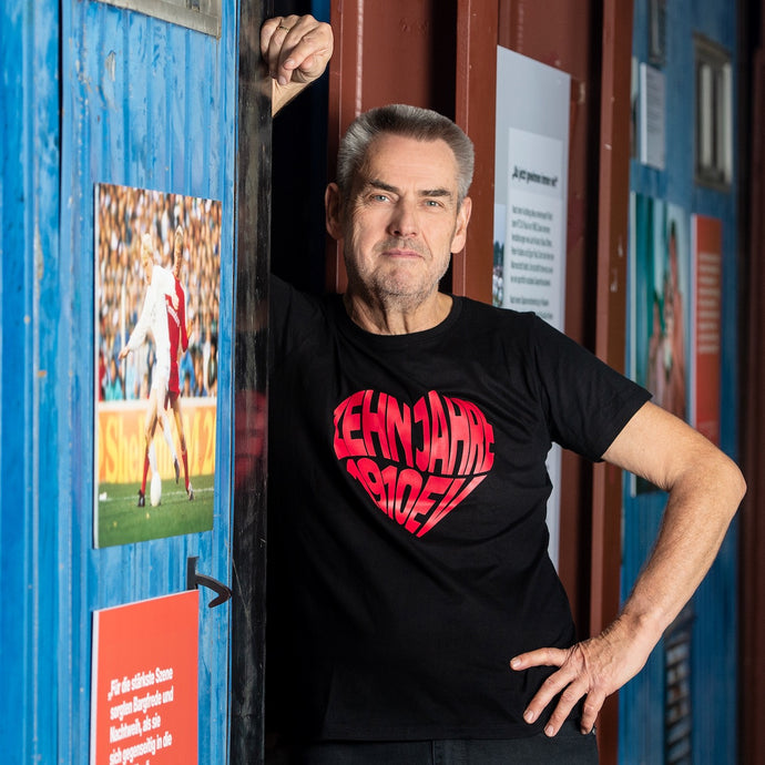 Auf dem Foto sieht man ein Model an eine blaue Wand gelehnt. Er trägt ein schwarzes T Shirt mit einem großen, roten Aufdruck  Zehn Jahre 1910 eV. Der Aufdruck ist herzförmig. Das Foto entstand im FC St.Pauli Museum.