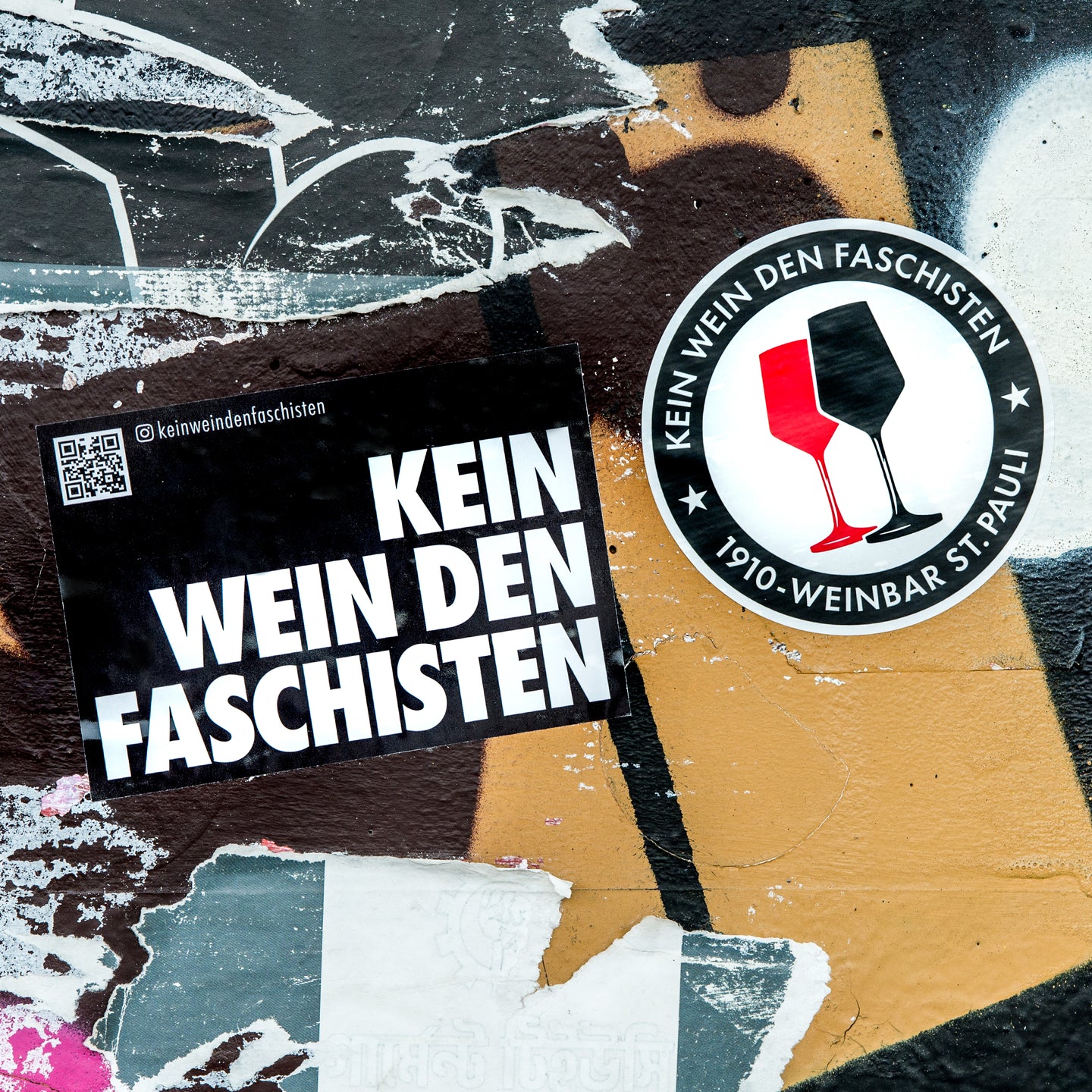 Auf dem Foto sieht man 2 Sticker an einer bunten Wand kleben. Der linke Sticker ist rechteckig und schwarz. In weißen Buchstaben steht darauf Kein Wein den Faschisten. Der rechte Sticker ist rund und es befindet sich kreisförmig der Schriftzug Kein Wein den Faschisten und 1910 Weinbar St.Pauli darauf. Sowie ein rotes und ein schwarzes Weinglas. In Anlehnung an das klassische Antifa Logo.