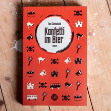 Lade das Bild in den Galerie-Viewer, Auf dem Bild sieht man einen Holzfußboden. Auf diesem liegt ein rotes Buch auf dem steht Toni Gottschalk Konfetti im Bier Roman. Es befinden sich außerdem viele, kleine, verschiedene Grafiken auf dem Cover. Diese sind schwarz und weiß. Zum Beispiel Totenköpfe, Busse, Trommeln, Fahnen und mehr.
