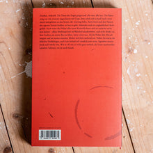 Lade das Bild in den Galerie-Viewer, Auf dem Bild sieht man einen Holzfußboden auf dem ein rotes Buch liegt. Es befindet sich ein kurzer Text darauf sowie ein schwarz weißer EAN Code. Es handelt sich um die Rückseite eines Buches.
