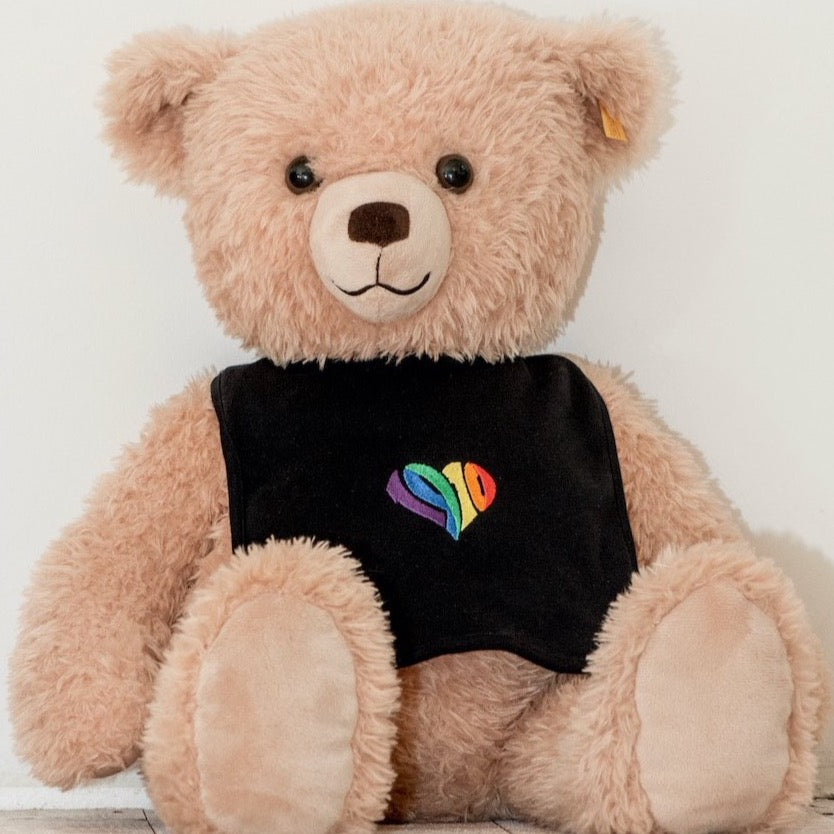 Auf dem Bild sieht man einen hellbraunen Teddybären. Dieser trägt einen schwarzen Babylatz. Auf dem Lätzchen ist mittig ein 1910 Herz in regenbogenfarben eingestickt.